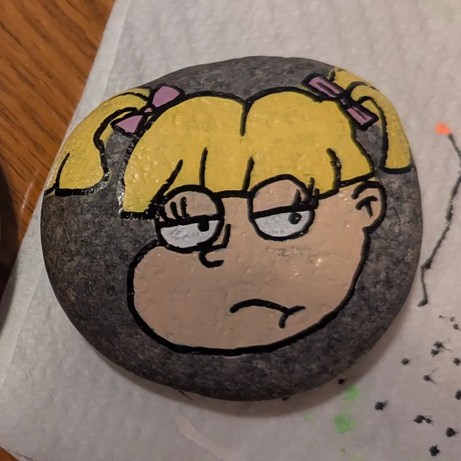 A round stone painted with Angelica from Rugrats, with blonde pigtails, purple bows, and a grumpy expression.
