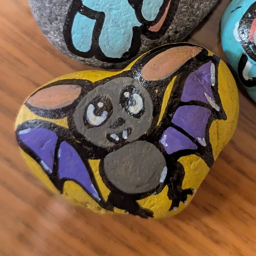 A stone painted with a round grey bat with big ears and purple wings against a golden-yellow background.