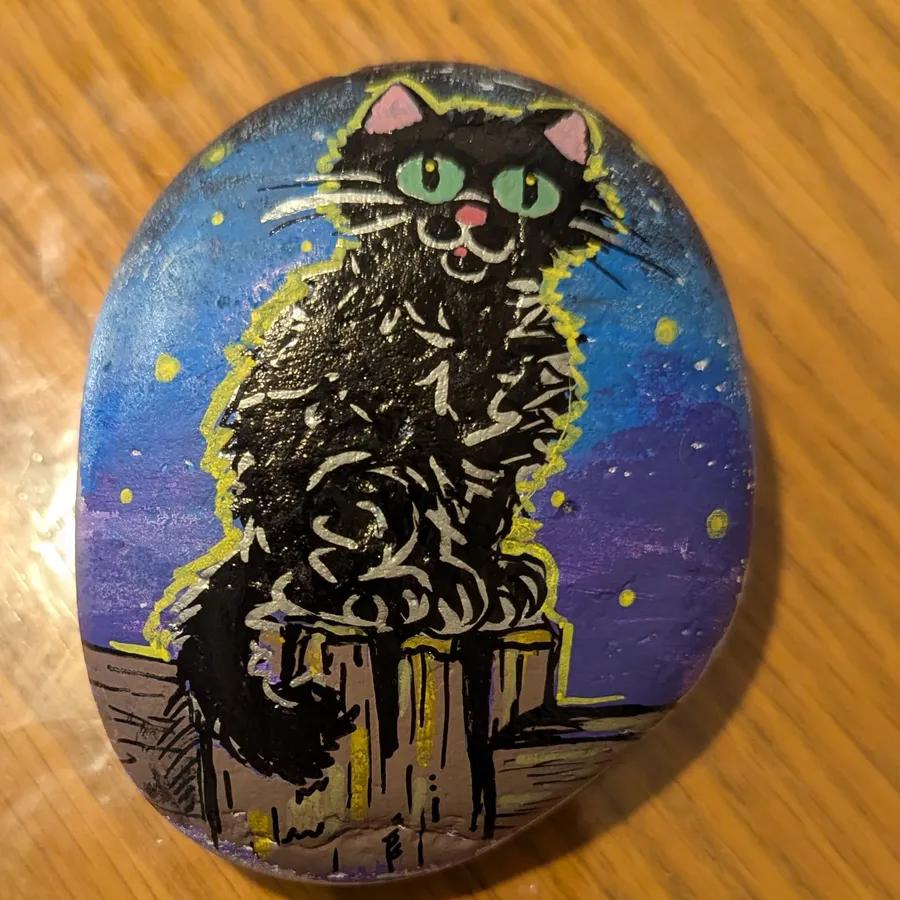 A stone painted with a scruffy black cat with green eyes, perched on a wooden post under a starry purple and blue night sky.
