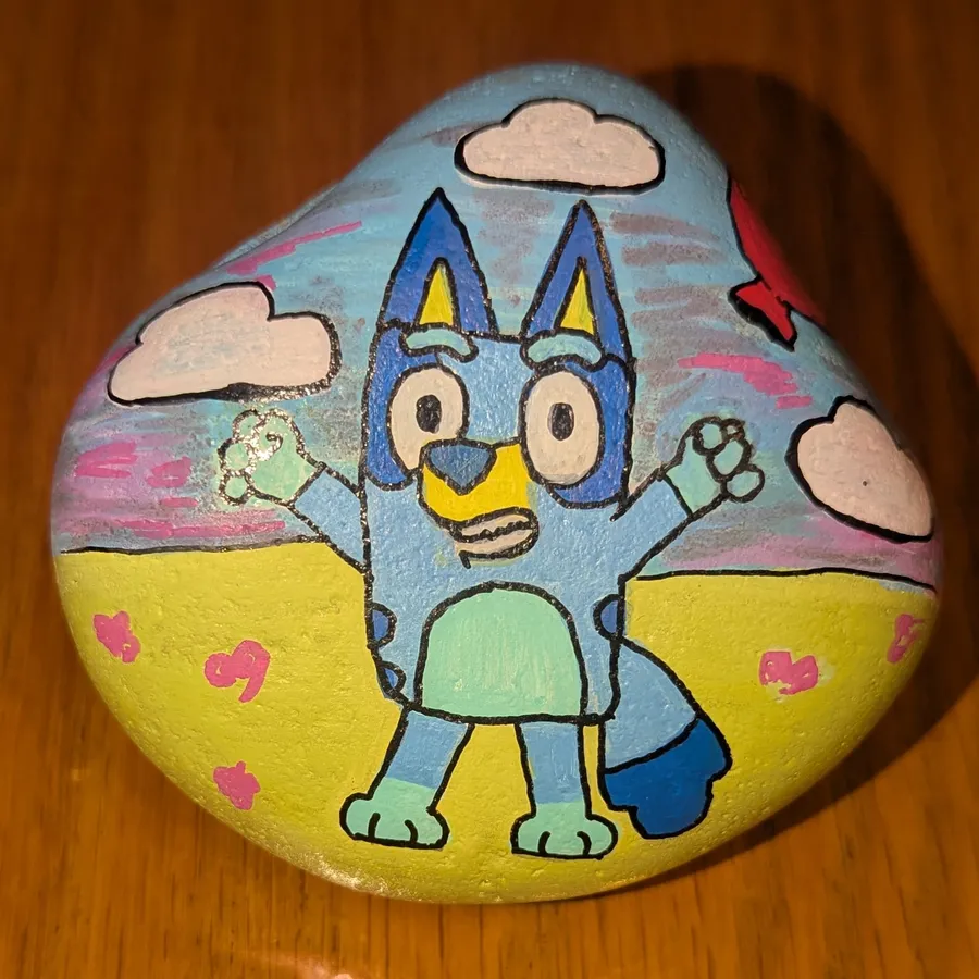 A stone painted with Bluey, the cartoon blue heeler puppy, waving under pink clouds on a yellow field.