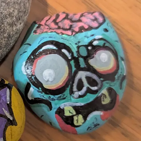 A stone painted as a teal zombie head with huge pale eyes, stitched teeth, and an exposed pink brain.