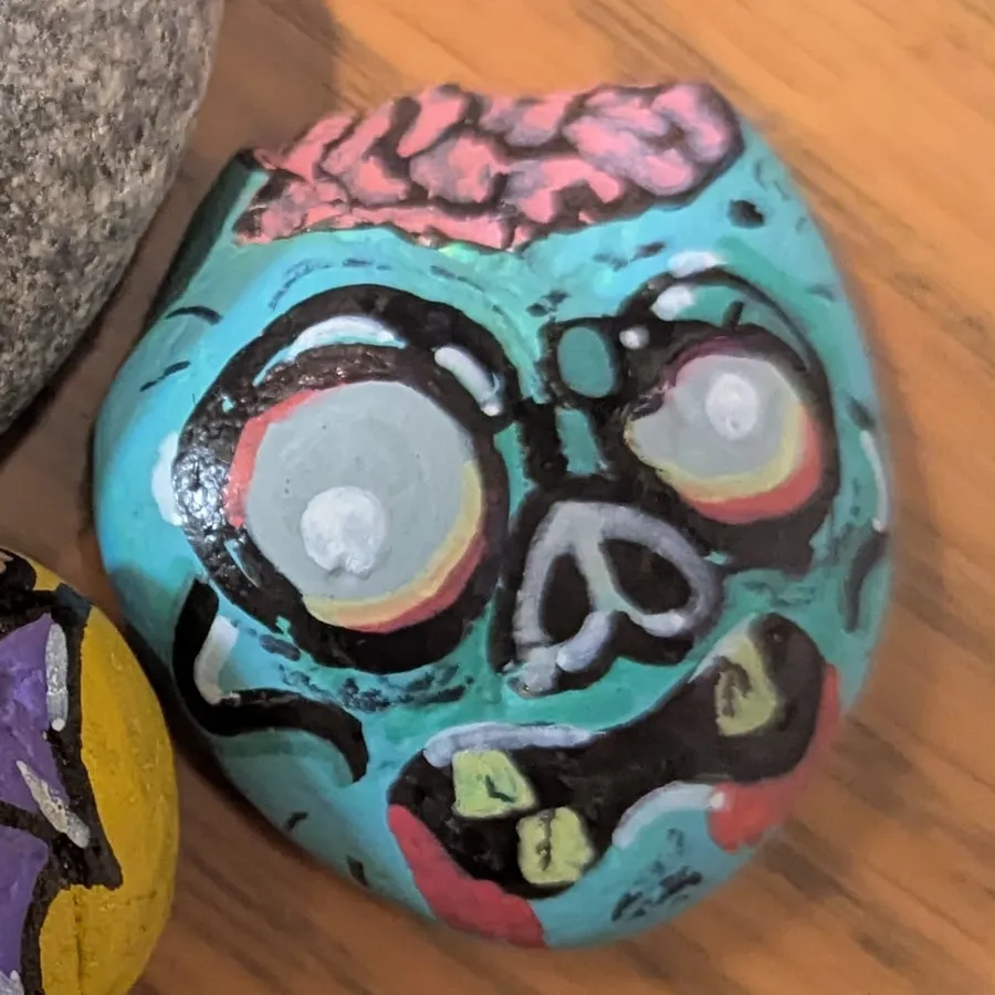 A stone painted as a teal zombie head with huge pale eyes, stitched teeth, and an exposed pink brain.