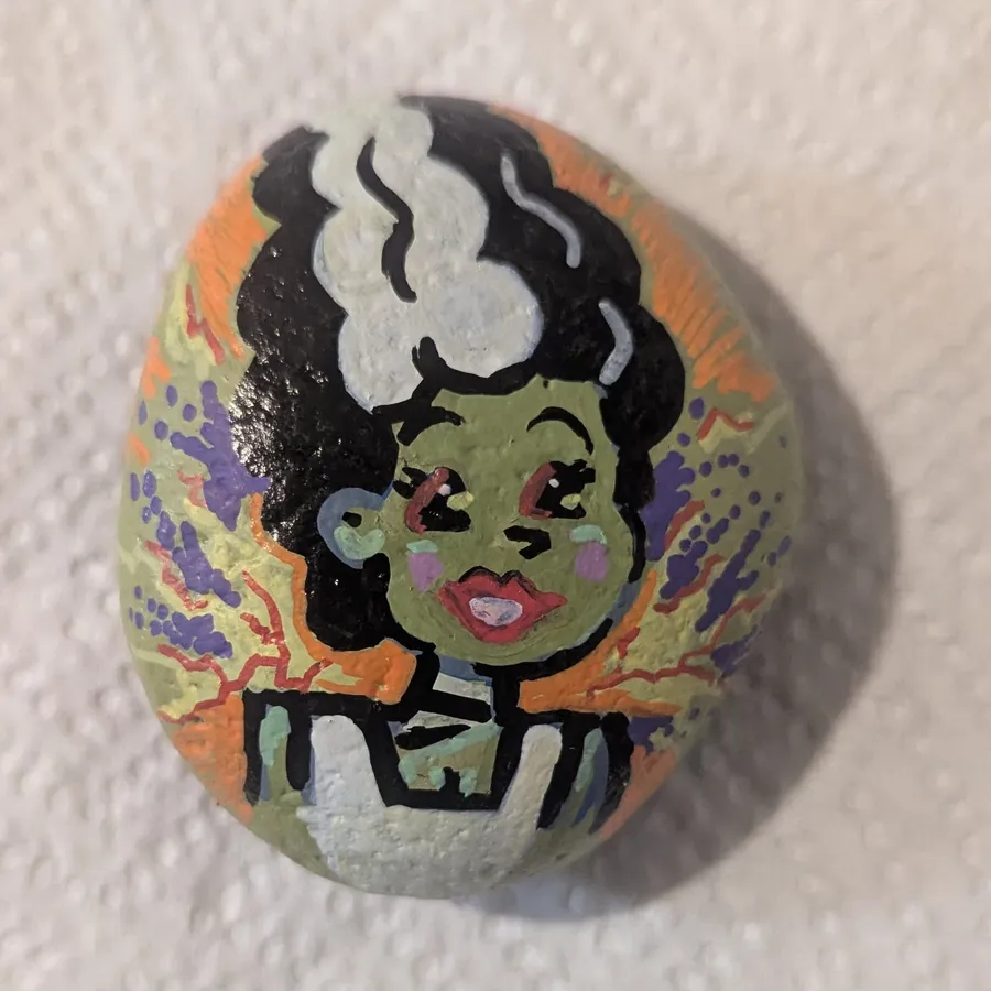 A stone painted with the Bride of Frankenstein, with her tall black-and-white streaked hair and purple lightning behind her.