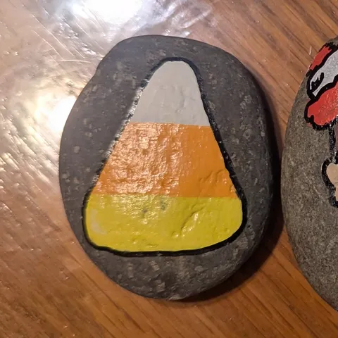 A dark river stone painted with a single piece of white, orange, and yellow candy corn.