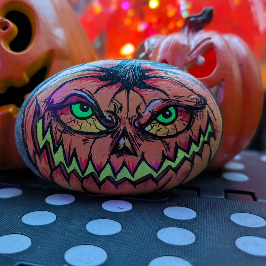 A smooth oval stone painted as a scowling jack-o'-lantern with glowing green eyes and a jagged neon-green grin.