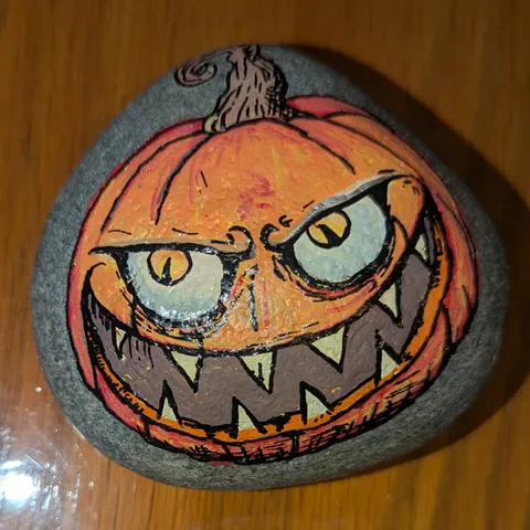 A grey stone painted as an orange jack-o'-lantern with a curling stem, pale glaring eyes, and a wide toothy grin.