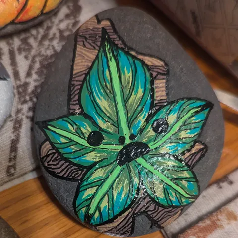 A dark stone painted with a teal and green leaf creature with a small sleepy face, set against carved bark.