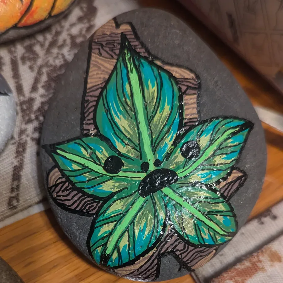 A dark stone painted with a teal and green leaf creature with a small sleepy face, set against carved bark.