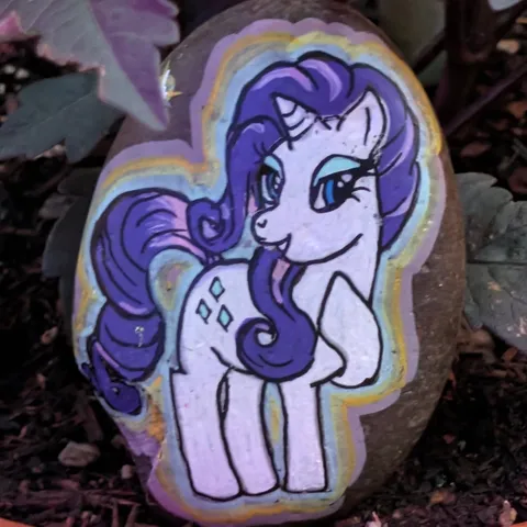 A stone painted with Rarity, the white unicorn with a purple mane from My Little Pony, outlined in a soft rainbow glow.