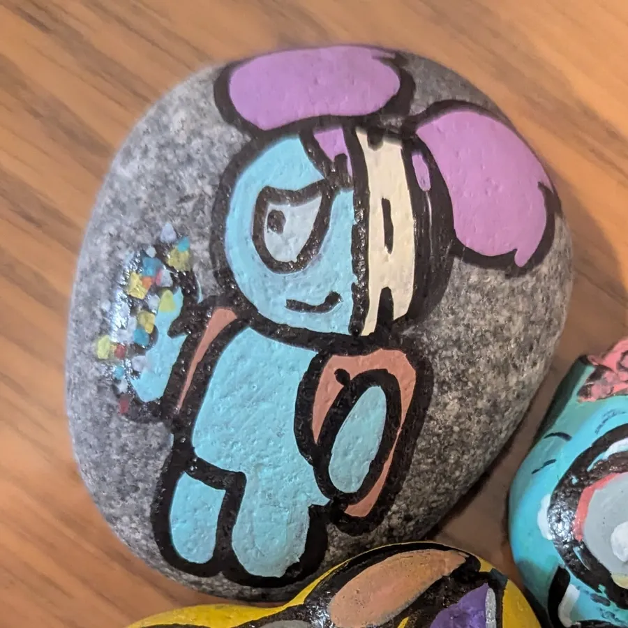 A grey stone painted with a pale blue, patched-up teddy bear wearing a pink bow and a bandage over one eye.