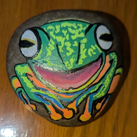 A dark stone painted with a grinning bright-green tree frog with bulging eyes and orange toes.